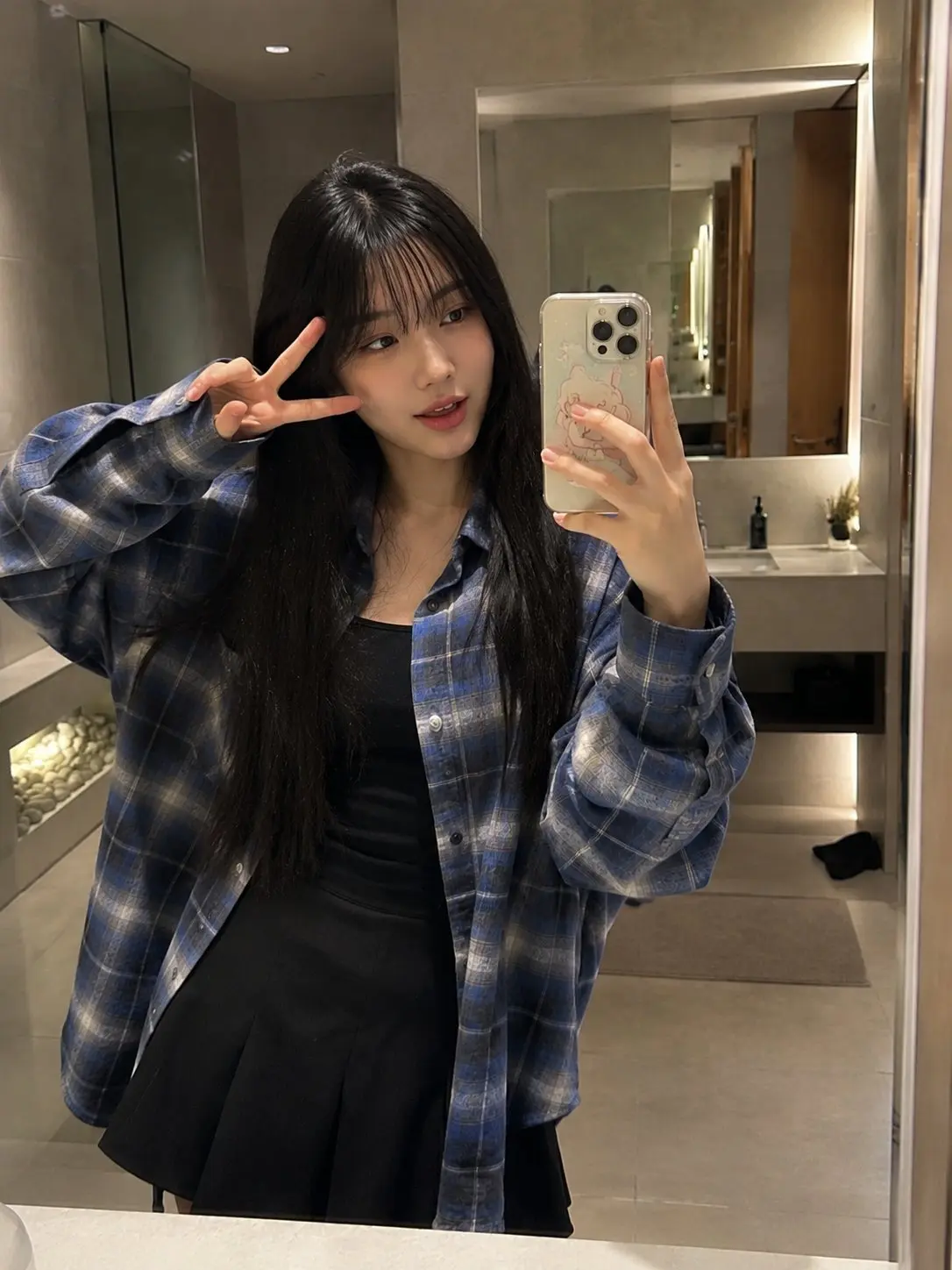 trend prompt: Beautiful young Japanese woman with long straight black hair and soft wispy bangs, wearing an oversized blue plaid flannel shirt over a black top and a modest black skirt. Standing in front of a bathroom mirror, holding a smartphone with a cute cartoon-style case, taking a casual mirror selfie. One hand lightly touching her hair, gentle smile and relaxed expression, natural everyday charm. Modern minimalist bathroom with neutral-toned tiles, large clean mirror, soft warm indoor lighting, contemporary sink and subtle decor visible in the background. Captured as an authentic handheld smartphone mirror selfie, realistic reflection, natural skin texture, slight phone-camera softness, casual social media lifestyle aesthetic, unedited everyday snapshot, imperfect framing, realistic iPhone camera quality, natural indoor shadows, cozy atmosphere, no professional photography setup, no studio lighting, no cinematic color grading, photorealistic, highly detailed, high-quality mobile photography, spontaneous everyday moment, realistic proportions, natural pose, wholesome and family-friendly content.