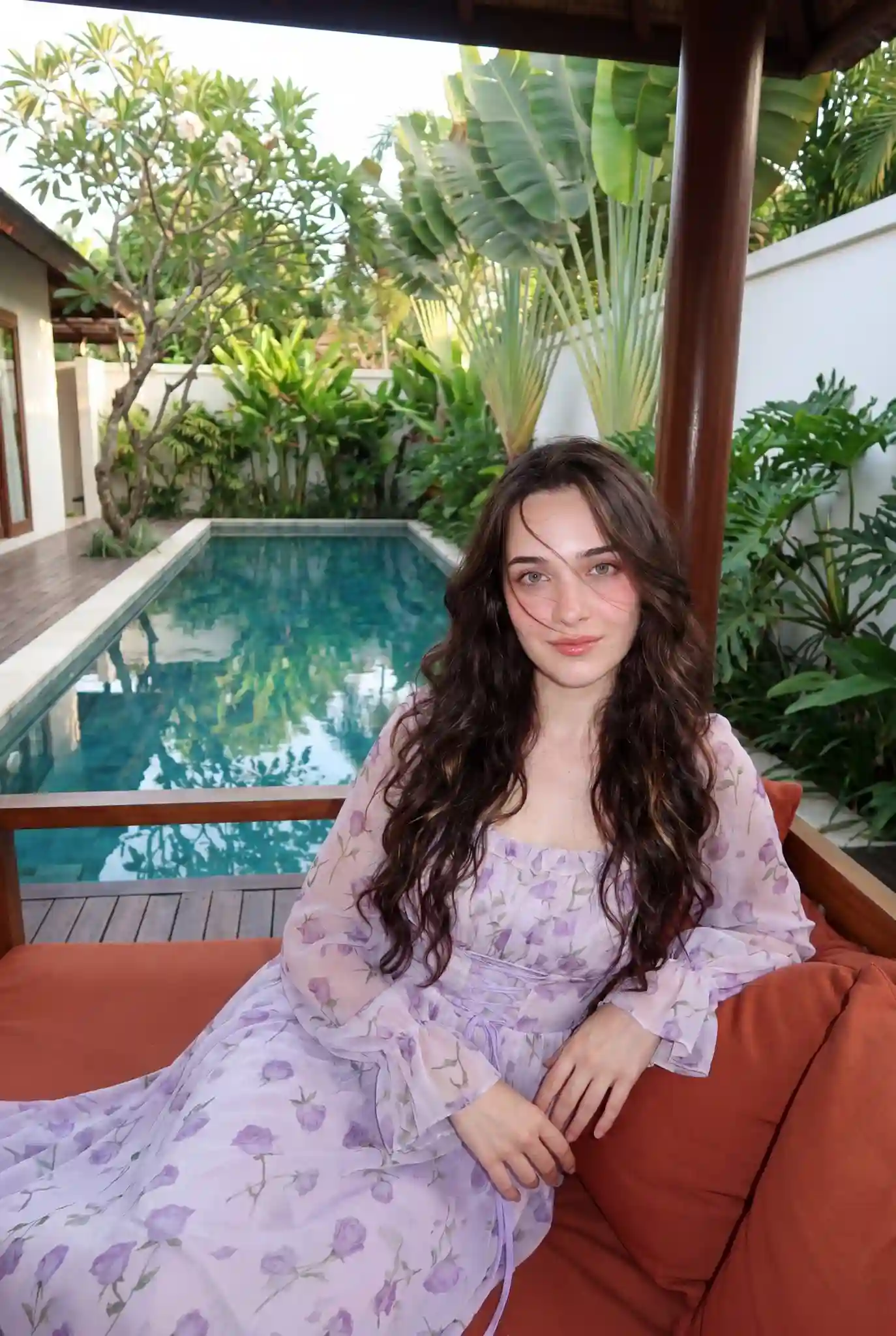 trend prompt: A highly realistic 8K photograph of a woman whose face matches the reference image. She has very fair, radiant, milky-white skin and a well-proportioned figure.
Hairstyle: Very long, thick, wavy hair with a glossy shine, featuring dark brown highlights. Slightly messy side part, with a few strands naturally blown across her face.
Face & Makeup: Dewy, glowing skin. Soft pink gradient blush applied on the cheeks and tip of the nose.
Lips: Glossy pink lips with smoothly blended edges.
Eyes: Light eyeliner, subtly shimmering eyeshadow, natural-looking lashes, and large almond-shaped eyes.
Outfit: Wears the outfit shown in the attached reference (image 2).
Pose: She is sitting in a pavilion, leaning back in an elegant and subtly sexy pose. Her hands rest comfortably. She looks toward the camera with a relaxed expression and a gentle smile.
Atmosphere: A peaceful, relaxing tropical garden setting  that blends nature with architecture.
Main elements in the scene:
Private Pool (Lap Pool): Clear blue water reflecting surrounding trees, creating a cool and refreshing mood.
Pavilion / Daybed: Located at the end of the pool, made of wood with brick-orange cushions, serving as a visual focal point and enhancing the relaxing vibe.
Natural Materials: Wooden decking and clean white walls that contrast beautifully with the green foliage and blue water.