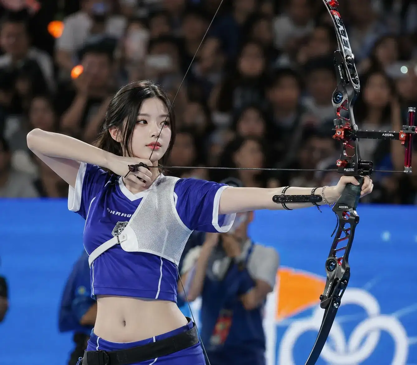 trend prompt: Ultra realistic sports photography, young East Asian female archer standing in a crowded indoor arena during a live competition, slim toned body, pale porcelain skin, delicate Korean visual beauty, soft oval face, straight black hair tied in a low ponytail with loose face-framing strands, calm confident expression, slight smile, focused elegant gaze. Natural Korean glass-skin makeup, smooth dewy skin, soft peach blush, subtle eyeliner, long lashes, gradient coral-red lips.
Wearing a fitted cropped royal-blue archery jersey with white trim, short sleeves, white athletic chest guard strapped diagonally across the torso, exposed waistline with toned abs, matching blue athletic skirt/pants with thin white stripe details, black waist belt, sporty professional archery styling.
Holding a modern black-and-red recurve bow vertically in the right hand with visible stabilizer and sight details, arrows gathered in left hand, relaxed elegant posture, body slightly angled, shoulders relaxed, standing confidently after a shot.
Background filled with blurred audience, photographers, colorful arena bokeh lights, live sports event atmosphere, teammates in matching blue uniforms softly out of focus. Cinematic indoor stadium lighting, soft spotlight on subject, realistic shadows, high contrast between subject and dark crowd, shallow depth of field, telephoto lens compression, natural motion blur in background, premium broadcast photography look, ultra detailed skin texture, realistic fabric folds, glossy lighting reflections, 85mm sports portrait lens, f/1.8, HDR, 8K, candid professional competition moment.