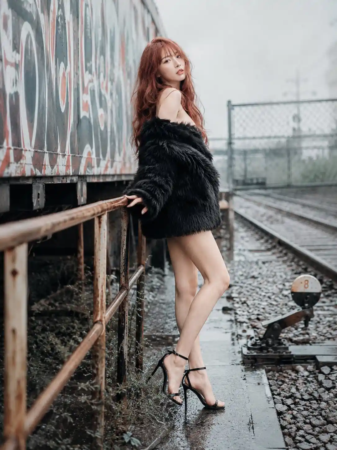 trend prompt: The image shows a 20-year-old adult East Asian woman with glamorous doll-like facial features, porcelain fair skin with a soft luminous glow, delicate oval heart-shaped face, clear warm hazel-brown eyes with glossy catchlights and long curled lashes, softly arched reddish-brown brows, small refined nose, peach-coral blush, warm champagne eye shadow, subtle eyeliner, glossy coral-rose lips with a softly parted pout, calm seductive fashion-editorial expression, long tousled copper-auburn red hair with darker natural roots, loose messy waves flowing past the chest and down the back, airy wispy see-through bangs falling softly across the forehead, face-framing side pieces and windswept layered texture, slim tall fashion-model body proportions with narrow waist, long legs, refined shoulders and collarbones, signature outfit locked as a black shaggy faux-fur off-shoulder mini coat worn as the main garment, plush high-pile texture, deep V neckline, bare shoulders and collarbones, oversized sleeves, mini-length hem, minimal black inner layer kept subtle under the coat, black ankle-strap stiletto sandals with thin straps and open toes, glamorous sunlit fashion portrait photographic realism, use the supplied character reference sheets as identity and outfit anchors, preserve the same face, hairstyle, body proportions, signature outfit, and overall character continuity while adapting only pose, expression, lighting, camera, and scene context, She is standing beside a railing. Let the image model choose a clearly varied non-default physically believable body arrangement within the selected pose base with distinct weight shift limb angles torso orientation and asymmetry compatible with the wardrobe camera framing and environment. Let the image model choose natural varied hand placement fitted to the selected body pose support contact wardrobe and camera crop without defaulting to stiff arms at the sides. Let the image model choose a natural head angle and orientation compatible with the camera angle body orientation and selected pose. The setting is graffiti-covered train yard edge, rusty railcar side, spray-painted metal surfaces, gravel track bed, rail switch hardware, chain-link fence fragment, weeds along the rails, rainy overcast environment, grey-blue rain sky, damp air, wet surfaces, muted visibility, cold daylight atmosphere, wet-surface reflected fill light on the subject, upward bounce from damp ground or walls, glossy reflected highlights on lower face and clothing. Inspired by Rinko Kawauchi, airy high-key image language. airy high-key photography, pale softly overexposed daylight glow, cool pastel tones, delicate everyday realism, fragile quiet details. The composition uses full body shot, full-length figure framing, wide framing, environmental scale, complete lower-body visibility, shoulder-level camera position, level lens axis near the shoulder line, stable upper-body portrait viewpoint, camera positioned on the subject's right side, 270-degree right profile view, lateral torso orientation, shot on 50mm standard lens, neutral perspective, minimal geometric distortion, natural subject proportions, balanced depth separation, moderate background compression, shallow depth of field, background defocus, controlled focus falloff behind the subject, clear subject plane separation, moderate optical blur gradient. high-key Japanese portrait color grade, elevated exposure, creamy porcelain highlights, softened shadow floor, restrained saturation, protected dark-material density, natural photographic detail, coherent subject construction, clear material readability, realistic spatial depth, do not add visible text unless explicitly requested.