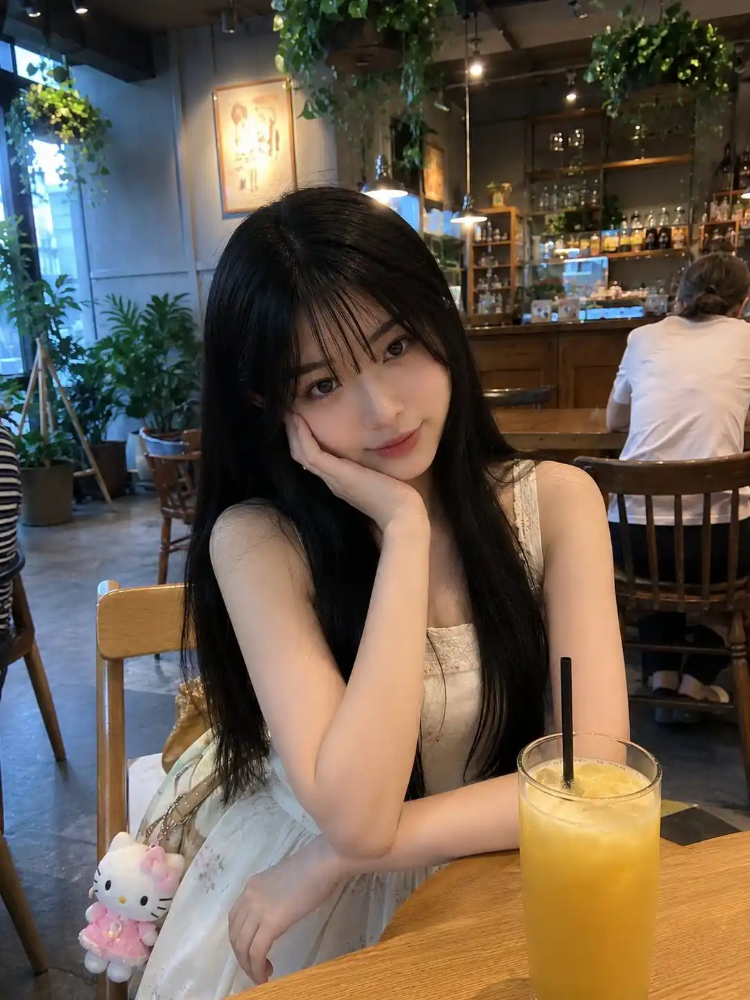 trend prompt: Young Japanese girl with long silky black hair, soft wispy bangs, fair glowing skin, and large expressive eyes, sitting casually at a cozy modern café. Wearing a sleeveless cream-white dress with delicate floral details, hands resting naturally in front of her, looking directly at the camera with a calm and slightly shy expression. A bright yellow fruit drink with a straw sits on the wooden table in the foreground. Cute Hello Kitty keychain hanging from her bag beside her chair. Warm café interior with hanging plants, large windows, decorative lighting, and soft ambient atmosphere in the background. Captured as a casual smartphone photo, handheld iPhone camera shot, natural indoor lighting, realistic skin texture, authentic mobile HDR processing, slight phone-camera softness, candid social media aesthetic, unedited everyday snapshot, natural colors, realistic shadows, ordinary café outing vibe, no professional photography setup, no studio lighting, no cinematic color grading, photorealistic, spontaneous lifestyle moment, high-quality mobile photography, cozy and charming everyday atmosphere.