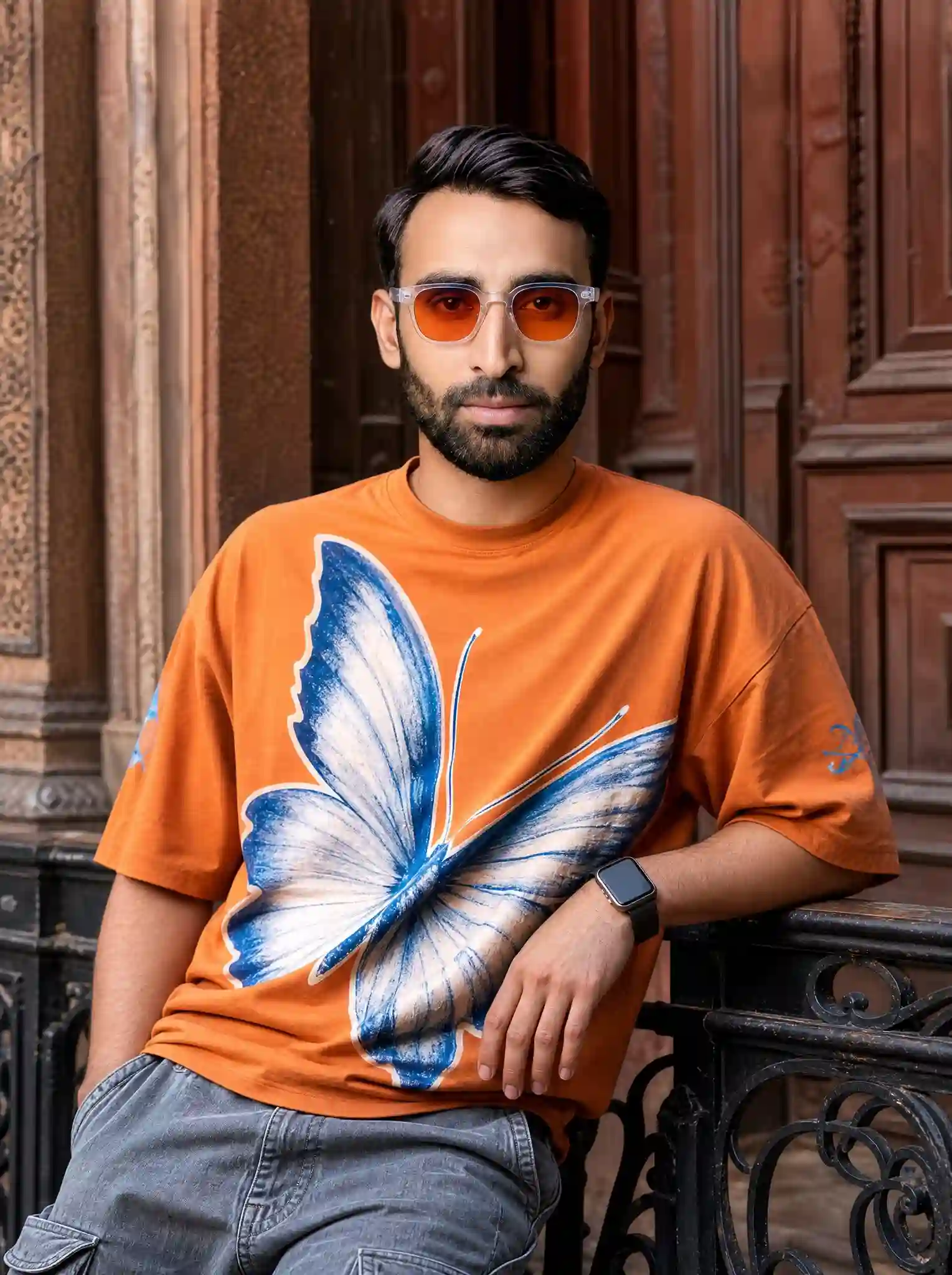 trend prompt: Use the uploaded image as the facial reference and preserve the exact facial identity with maximum accuracy. Maintain the same face structure, eye shape, eyebrows, nose, lips, jawline, skin tone, hairstyle characteristics, beard/facial hair details (if present), and all recognizable facial features from the reference image. Ultra-realistic medium shot portrait of the same young South Asian man leaning casually against a dark, ornate wrought-iron railing with a confident expression and full attitude. He is wearing an oversized vibrant orange t-shirt featuring a large detailed white and blue butterfly graphic on the front, paired with stylish grey denim cargo pants and a modern smartwatch. His hairstyle is stylish and natural, with well-groomed masculine appearance. He is wearing orange gradient sunglasses with a white transparent frame. BACKGROUND: A rustic textured reddish-brown wooden door behind him, vintage architectural details, realistic outdoor setting, cinematic atmosphere. POSE & COMPOSITION: Medium shot, relaxed confident pose, masculine fashion editorial style, casual streetwear look, natural body posture, premium fashion photography composition. CAMERA & LIGHTING: Shot on an 85mm lens, DSLR quality, shallow depth of field, warm cinematic lighting, soft natural daylight, realistic shadows, professional photography look. STYLE: Hyper-realistic, photorealistic, ultra detailed, 8K resolution, HDR, realistic skin texture, sharp focus, natural colors, luxury fashion magazine quality, cinematic depth, highly detailed fabric texture, realistic masculine features. Aspect Ratio: 4:5 Vertical Negative Prompt: Low quality, blurry, cartoon, illustration, painting, fake face, different person, changed identity, distorted face, bad anatomy, extra fingers, extra limbs, deformed hands, unrealistic body, plastic skin, oversaturated colors, watermark, logo, text, noise, artifacts.