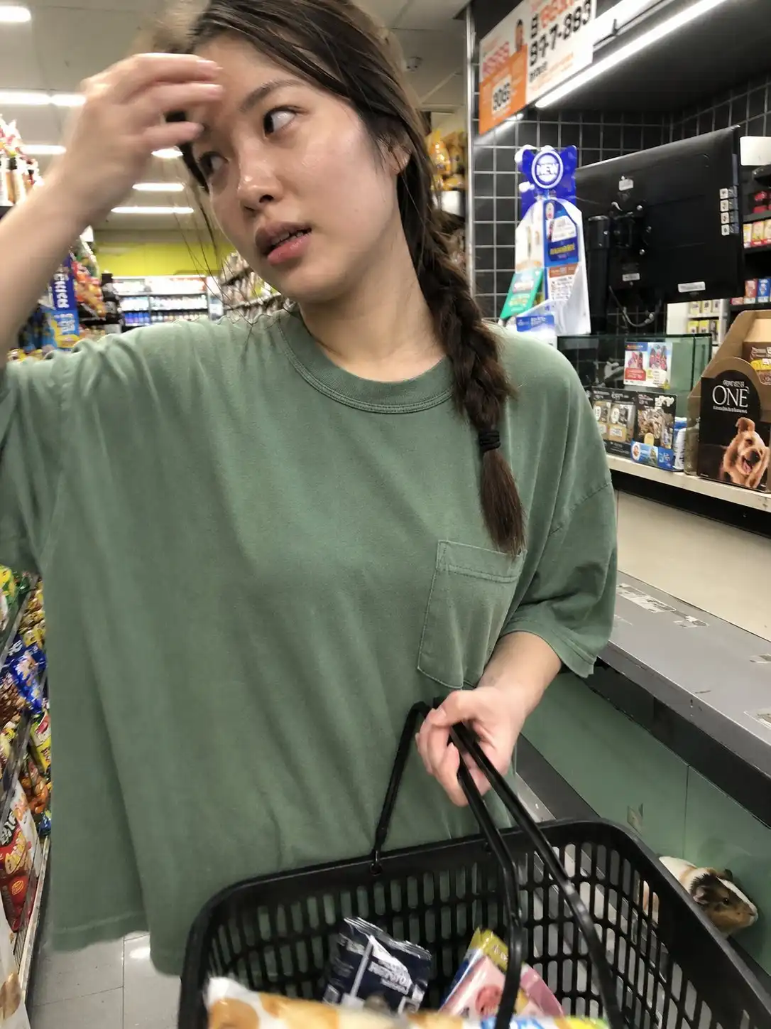 trend prompt: Accidental candid iPhone front-camera photo captured inside a small neighborhood convenience store. A young Korean woman stands in the checkout line, casually wiping a trace of sweat from her forehead while balancing a shopping basket. She looks off to the side with a slightly impatient, distracted expression, completely unaware of the camera. Faded sage-green oversized cotton T-shirt, relaxed fit, loose braid draped over one shoulder. Natural skin texture with visible pores, subtle facial shine from indoor warmth, realistic imperfections, no beauty retouching. Bright overhead fluorescent lighting creates cool-toned shadows and a faint yellow-green store color cast. Authentic smartphone image quality from an older iPhone, slight motion blur on the moving hand, mild sensor noise, compressed detail, imperfect framing, spontaneous everyday moment. Cluttered convenience-store aisles and checkout displays in the background. A small guinea pig is barely visible near the bottom corner of the frame, appearing accidentally included in the shot.