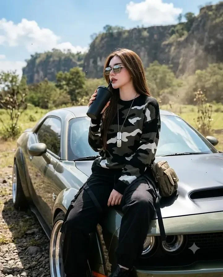trend prompt: Photorealistic 3/4 body portrait taken with a Nikon D780, 80mm lens, f/3.4, high-angle camera from the side. A beautiful young woman leans casually against a modified Dodge Challenger, sitting on the car's hood with her legs crossed and feet resting naturally below. She looks off into the distance with a cool, relaxed expression. Her left hand holds a matte black tumbler with a lid, raised naturally toward her face as if taking a sip. A stylish crossbody pouch bag rests beside her. She has healthy, natural skin with realistic texture, long black hair with soft natural volume, and subtle makeup that enhances her features while maintaining a realistic appearance. She wears an oversized black knit sweater with a camouflage pattern in dark gray, white, and army green, paired with a silver military-style nameplate necklace. She also wears slightly loose black tactical cargo pants with hanging straps in a streetwear style, creating an edgy and fashionable look. She has silver-gray reflective box-frame sunglasses. The background features a slightly dry green meadow with scattered plants and trees, drifting sand dust, towering rocky cliffs, and a bright blue sky with clearly visible white clouds. The scene has the feel of cool cinematic street photography with a slightly shallow depth of field, dramatic shadows, gritty film texture, ultra-realistic details, 8K quality, and a slightly bright yet cool color tone. Light enters from the right, creating a beautiful cinematic lens flare reflected on the car's glass. Aspect ratio: 4:5.