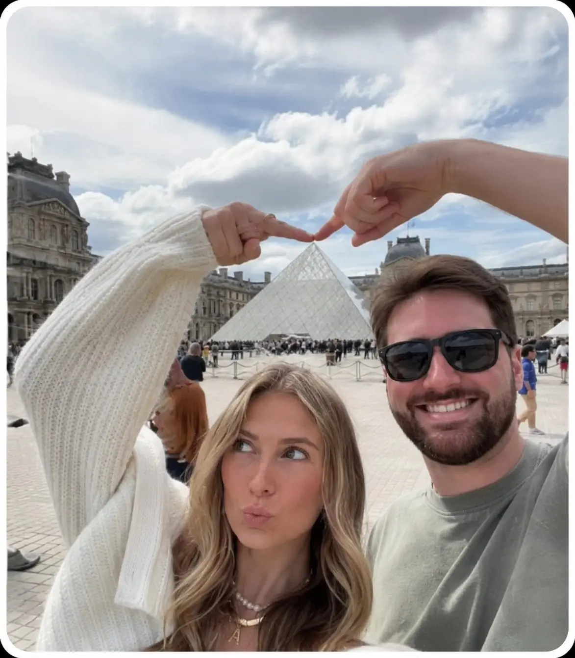 trend prompt: Ultra-realistic travel selfie of a happy young couple standing in front of the Louvre Pyramid in Paris. The woman has long wavy blonde hair, wearing an oversized chunky white knitted cardigan with extra-long sleeves, layered pearl and gold necklaces including a gold letter "A" pendant, making a playful duck-face expression while looking slightly upward. The man has short brown hair, a full trimmed beard, black sunglasses, a warm smile, wearing an olive-green t-shirt. They recreate the famous "Creation of Adam" pose by touching their index fingers together directly above the tip of the Louvre glass pyramid, creating a perfect visual illusion. The woman raises her left arm, the man raises his right arm. Both are framed in a close-up selfie composition. Background features the Louvre Museum courtyard in Paris with the iconic glass pyramid centered, historic palace architecture, tourists walking in the distance, dramatic blue sky with large white clouds, bright natural daylight, realistic travel photography, soft natural colors, ultra-detailed skin texture, DSLR quality, 35mm lens, HDR, sharp focus, cinematic composition, Instagram travel aesthetic. White rounded photo border around the entire image, clean composition, no text, no watermark, no logo, no collage, no inset image, no clay style, no cartoon, no illustration, photorealistic, 8K, masterpiece, best quality.