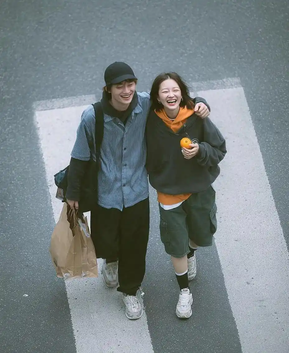 trend prompt: A cinematic overhead street photo of a young couple walking on a zebra crossing, candid and natural interaction, boy happily laughing with her arm over the girl's shoulder, playful romantic atmosphere. Boy wearing a black cap, oversized pale blue shirt, dark sweater around shoulders, loose black pants, sneakers, carrying a brown paper shopping bag. Girl wearing an oversized dark sweatshirt with an orange sweater tied over her shoulders, green shorts, black socks and white sneakers, holding a small orange fruit. Soft daylight, muted color grading, realistic skin texture, documentary photography, subtle film grain, Korean/Japanese lifestyle aesthetic, emotional storytelling, high-angle top-down composition, natural movement, ultra realistic, masterpiece.