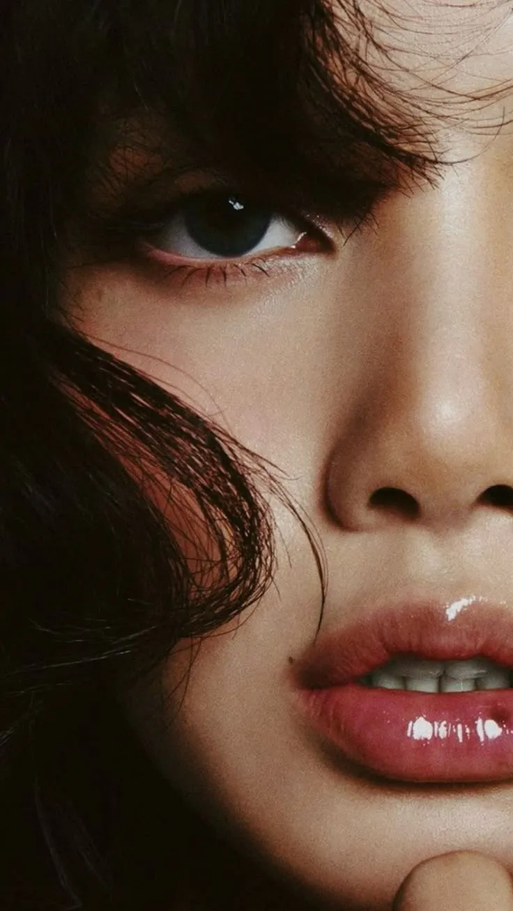 trend prompt: An ultra-realistic editorial beauty portrait featuring an exceptionally attractive young East Asian woman captured in an extreme close-up macro composition. Only the upper half of the face and lips are visible, creating an intimate, cinematic framing. One large almond-shaped dark brown eye is sharply in focus while the second eye is naturally concealed behind soft, tousled jet-black curtain bangs. Long, delicate eyelashes with subtle eyeliner and natural eye makeup enhance the captivating gaze. Her complexion is porcelain-fair with smooth, flawless skin displaying natural pores and subtle skin texture. The lighting softly sculpts the face, highlighting the bridge of the nose with gentle specular reflections while preserving realistic shadows. A tiny beauty mark sits near the left side of the upper lip, adding character. Her lips are full, plump, and slightly parted, revealing a glimpse of perfectly aligned white teeth. They are coated with a thick, high-shine transparent gloss, producing bright reflective highlights and a wet, glass-like finish. Lip color is a soft natural pink with smooth gradients and realistic texture. Messy, layered black hair frames the face, with loose strands flowing across the forehead and cheek, creating depth and organic movement. Individual hair strands remain crisp and highly detailed. A single curled lock of hair rests against the cheek, adding softness and visual balance. The expression is calm, mysterious, and subtly seductive, with relaxed facial muscles and a slightly open mouth. One fingertip gently touches beneath the chin, barely entering the frame, creating a natural beauty editorial pose. Lighting consists of soft directional studio light from the upper right, producing cinematic shadows, delicate skin highlights, and rich contrast without harsh clipping. The color grading is warm, muted, and slightly desaturated with deep blacks, soft beige skin tones, and glossy highlights, resembling luxury fashion magazine photography. The background is entirely filled by hair and facial features, with no distractions. Extremely shallow depth of field isolates the eye and lips while maintaining soft falloff across the frame. Style: luxury beauty editorial, Vogue, Harper's Bazaar, cinematic portrait, macro beauty photography, ultra photorealistic, premium skincare campaign, fashion magazine cover, fine art portrait. Camera: Sony A7R V, 90mm macro lens, f/2.0, ISO 100, 1/200s, ultra-high resolution, HDR, razor-sharp eye focus, natural skin texture, realistic hair strands, professional studio lighting. Quality Tags: masterpiece, best quality, 16K UHD, hyperrealistic, photorealistic, RAW photo, ultra detailed, pore-level skin texture, cinematic lighting, global illumination, ray tracing, HDR, high dynamic range, natural color science, crisp focus, extremely detailed eyes, glossy lips, premium editorial photography.
