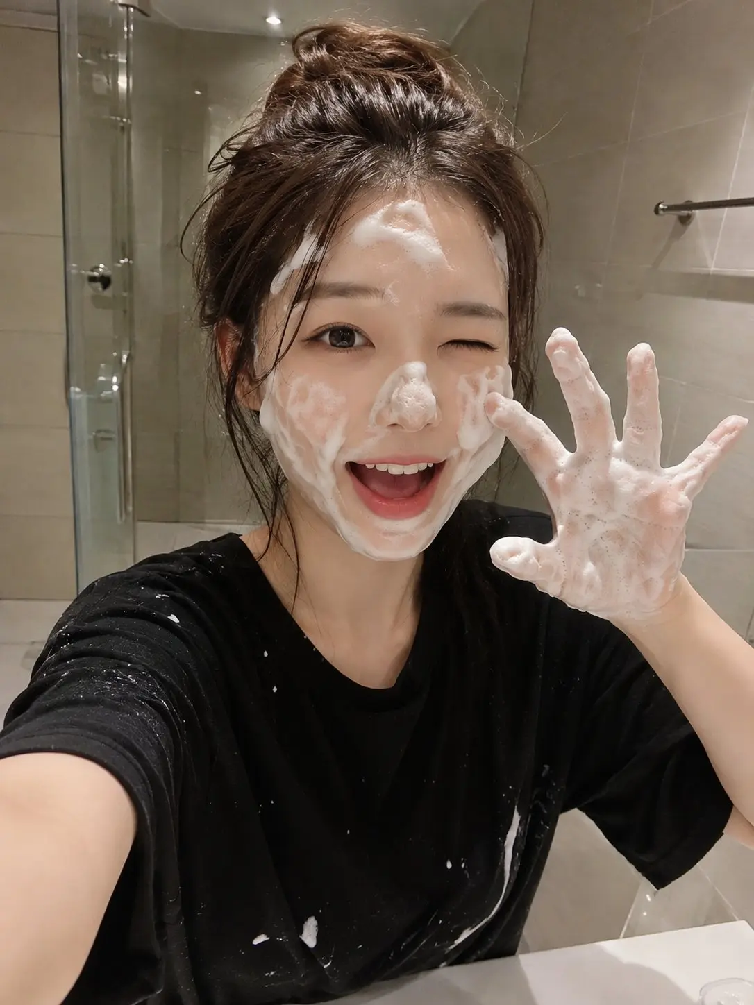 trend prompt: A RAW ultra-realistic smartphone photo of a cute young Japanese woman in a modern bathroom during her skincare routine. She has fair porcelain skin, large expressive dark brown eyes, naturally soft pink lips, and long chestnut-brown hair that is slightly damp and tousled. She is playfully winking with one eye while smiling with her mouth open, both hands raised in a cute claw pose completely covered in rich white facial cleansing foam. Thick soap bubbles are spread naturally across her forehead, cheeks, nose, chin, hairline, and fingers. She wears a loose oversized black T-shirt with a few soap splashes on it. The background features a clean minimalist bathroom with beige ceramic wall tiles, a frameless glass shower door, a chrome towel bar, and a white sink. The image is captured as a casual selfie using a modern smartphone held at arm's length, creating an authentic everyday social media aesthetic. Warm indoor bathroom lighting, realistic skin pores, subtle smartphone HDR, natural shadows, tiny sensor noise, automatic white balance, slight handheld framing, 26mm smartphone lens, realistic exposure, no studio lighting, no beauty filters, no AI-generated appearance, ultra-photorealistic, indistinguishable from a real iPhone photo, RAW mobile camera quality, 8K.