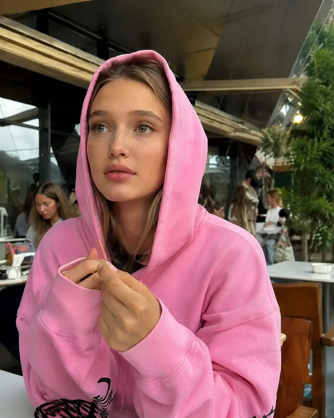 trend prompt: Ultra-realistic candid portrait of a beautiful young woman sitting at an outdoor café, wearing an oversized pastel pink hoodie with the hood up. She has fair skin, soft light-brown hair partially visible under the hood, natural makeup, large expressive blue-green eyes, and a calm, thoughtful expression while looking slightly upward. She is gently holding a thin wooden coffee stirrer between her fingers close to her chest. Modern street café setting with glass walls, metal canopy, wooden chairs and tables, blurred customers in the background, green trees and soft natural daylight. Cozy urban atmosphere with shallow depth of field and cinematic bokeh. Photorealistic DSLR photography, 85mm lens, f/1.8, HDR, ultra-detailed skin texture, realistic fabric folds, natural lighting, soft color grading, high dynamic range, premium editorial lifestyle photography, Instagram aesthetic, masterpiece, best quality, ultra sharp focus, 8K. Negative prompt: cartoon, anime, CGI, clay, 3D, painting, illustration, low quality, blurry, noisy, watermark, logo, text, frame, border, collage, duplicate person, deformed hands, extra fingers, distorted face, cropped head.