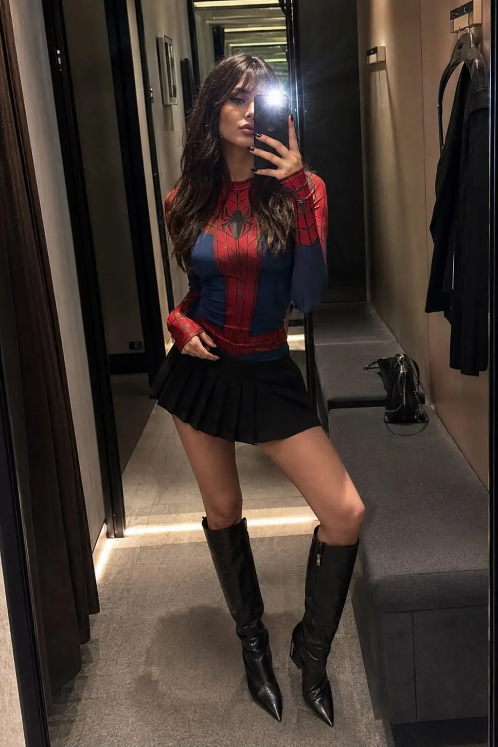 trend prompt: Use the uploaded reference image as the only identity reference. Preserve her facial features, face shape, skin tone, body proportions, hairstyle, and overall appearance exactly as in the reference. Create an ultra-photorealistic mirror selfie of the same young woman inside a stylish boutique fitting room with warm ambient lighting and elegant full-length mirrors. She is wearing a form-fitting red-and-blue spider-inspired long-sleeve top paired with a sleek black pleated mini skirt and knee-high black leather boots, creating a fashionable superhero-inspired look rather than a costume. Her long, straight dark hair with soft bangs falls naturally over her shoulders. She holds her smartphone in front of her face with the camera flash on, partially obscuring her features in a realistic mirror-selfie style. The fitting room features a cozy modern interior with textured walls, a bench, subtle reflections, and soft cinematic shadows. Capture lifelike skin texture with visible pores, realistic fabric details, natural body proportions, shallow depth of field, editorial fashion photography styling, ultra-detailed realism, and crisp 4K quality.