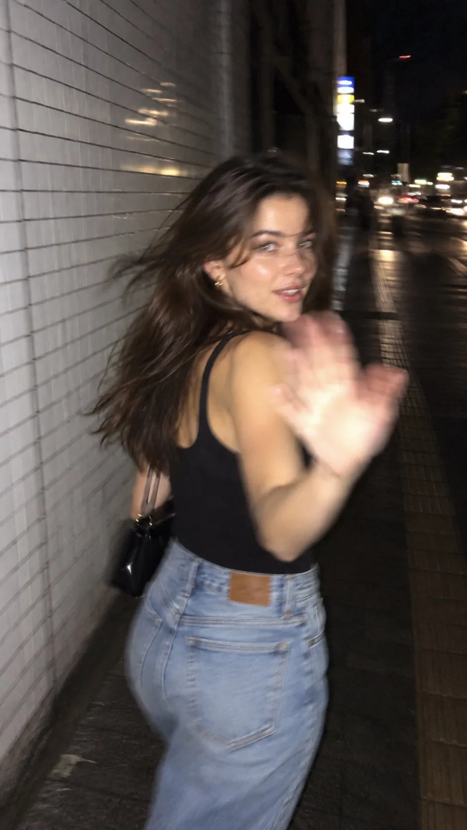 trend prompt: Ultra-realistic late-night smartphone snapshot, 9:16 vertical. Use the reference only to establish the same fictional female character's overall appearance and continuity. Keep her recognizable across the series with the same dark-brown wavy hair, natural features, black fitted sleeveless tank top, loose high-waisted light-blue jeans, small gold hoop earrings, delicate necklace, white sneakers, and small black shoulder bag. Scene: A quiet Tokyo side street late at night after light rain. She walks beside an off-white ceramic-tiled building while distant convenience-store lights, traffic signals, parked bicycles and small Japanese storefronts softly disappear into the darkness. Wet pavement catches scattered reflections. New candid moment: Photograph her from a slightly low rear three-quarter angle, about two steps behind her. She has just slowed down and casually turns only her upper body toward the camera while continuing forward. One hand is loosely tucking windblown hair behind her ear while the other hangs naturally beside her bag. She looks toward the camera from the corner of her eyes with a tiny closed-mouth smile, as though she noticed her friend taking another photo and is trying not to laugh. No deliberate fashion pose. No exaggerated expression. Her shoulders remain relaxed, hips and legs naturally positioned from walking, with one foot caught halfway through the next step. Camera: Modern smartphone rear camera with direct flash. Photographer walking at the same time, creating subtle accidental camera shake. Slight motion blur around her hair, moving leg and fingertips, while her eyes remain just barely readable. Mild rolling-shutter distortion, imperfect autofocus, slight edge softness and realistic digital noise. Composition should feel accidental: her body sits slightly off-center, a little too much empty darkness on one side, with part of her shoe approaching the bottom edge of frame. Flash & exposure: Hard direct phone flash illuminates her face, shoulder, black top and denim while the street behind her falls rapidly into darkness. Slightly overexposed skin highlights, hard natural shadow against the tiled wall, shiny flash reflections on individual hair strands and subtle denim texture. Distant lights bloom and smear naturally. Aesthetic: Raw Pinterest night-photo-dump energy. Intimate, youthful, spontaneous and imperfect. It should look like a friend took the photo during a random Tokyo night walk-not a photographer conducting a photoshoot. Real pores, tiny skin variations, flyaway hairs, natural fabric wrinkles, realistic body proportions and ordinary smartphone processing. Absolutely no: studio lighting, cinematic teal-orange grading, professional bokeh, fashion-editorial posing, perfect symmetry, airbrushed skin, artificial beauty retouching, HDR look, DSLR sharpness, staged composition, AI-perfect anatomy or polished commercial photography. The final result should feel like an unexpectedly beautiful photo hidden among 30 messy pictures from the same night