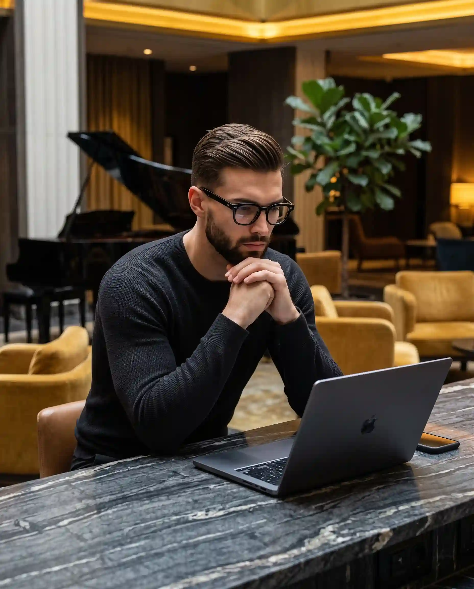 trend prompt: A focused young adult male sits leaning forward at a counter, wearing a fitted dark grey-black knit long-sleeve shirt and thick-rimmed black glasses. His short-to-medium brown hair reaches the top of his ears in a classic slicked-back style, featuring a natural straight to slightly wavy texture with visible separation, subtle flyaways, and uneven sections on the sides, styled with moderate hold product for a glossy wet look, brushed straight back with moderate volume at the front quiff. His expression is genuinely serious and calm as he looks down at an open dark grey aluminum laptop. His slightly hunched, relaxed shoulders lead down to his elbows resting on the counter, supporting his weight as his hands are loosely clasped together in front of his lower face and chin, fingers organically interlaced in deep thought. He is positioned centrally at a dark grey stone counter characterized by subtle white veining, where a smartphone in a dark case rests flat beside the laptop. The deep, luxurious background reveals a high-end hotel lobby or lounge, featuring a pristine high-gloss black grand piano with its lid down and a large leafy green Ficus plant near a window in the left midground, balanced by plush, curved mustard yellow armchairs and a glowing conical table lamp on the right. The walls are a smooth, matte warm beige and taupe, accented by tactile vertical ribbed dark brown wood paneling and softly illuminated framed artwork over dark brown solid wood plank floors. The moody, sophisticated atmosphere is bathed in warm golden artificial lighting from multiple overhead recessed spotlights and practical background lamps, creating a cinematic medium contrast. Soft, gradual gray shadows pool under his chin, beneath his arms, and below the laptop on the counter, while preserved highlights gently illuminate his forehead, cheekbones, glasses frames, and glossy hair. The analogous warm palette blends dark charcoal, dark brown, taupe, and striking mustard yellow accents. Captured with digital photography styling a realistic dark academia aesthetic using a 50mm lens at f/2.8, 1/60s, and ISO 800 with a warm white balance, the image boasts a medium depth of field that keeps the subject in sharp focus while rendering the rich background in a soft, smooth bokeh with subtle digital noise in the shadows, finished with warm color grading, lifted blacks, and a soft vignette, all beautifully framed in a 4:5 aspect ratio