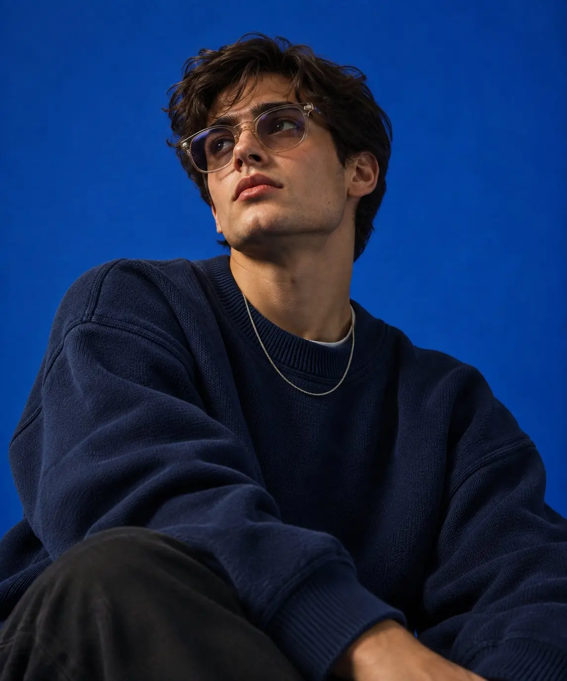 trend prompt: Create an ultra-photorealistic high-end studio fashion portrait of a handsome young adult man in his early-to-mid 20s, photographed like a luxury streetwear campaign. Subject & Appearance: A naturally handsome young man with masculine yet refined facial features, realistic skin texture, subtle natural imperfections, expressive eyes, defined jawline, straight nose, natural lips, and light authentic stubble. Tousled, slightly wavy dark-brown hair with realistic individual strands. His appearance should feel believable and photographic rather than overly perfected or AI-generated. Pose & Expression: Place him in a relaxed seated pose, leaning slightly toward the camera. His torso is angled subtly while his shoulders remain relaxed. One arm is bent upward with his hand resting naturally near his mouth and chin, with his fingers positioned loosely and realistically. His other arm rests casually in the foreground. His head is tilted slightly upward and toward the side, with a calm, confident, slightly aloof expression. His gaze is directed toward the camera through the glasses. The pose should feel effortless and editorial rather than staged. Wardrobe: Dress him in a premium oversized dark-navy textured knit sweater with a relaxed streetwear silhouette. The sweater should have realistic heavy fabric, natural folds, ribbed cuffs, and believable draping around the shoulders and arms. Pair it with relaxed-fit light-wash blue denim jeans. Add a minimal fine silver chain necklace. Keep accessories understated and luxurious. Eyewear: Add sophisticated oversized translucent/clear-frame glasses with lightly tinted smoky lenses. The glasses must have realistic reflections and accurate interaction with the studio lighting. Ensure the frames sit naturally on the face without distorting the eyes or facial proportions. Studio & Background: Use a completely seamless, rich cobalt-blue studio backdrop extending uniformly across the entire frame. No visible studio equipment, furniture, texture, gradients, patterns, or environmental distractions. Maintain a premium monochromatic blue fashion-campaign aesthetic. Lighting: Use soft directional studio lighting coming from slightly above and to one side of the subject. Create subtle highlights across the forehead, cheekbones, nose, lips, glasses, and hair while maintaining realistic shadow transitions along the jawline, neck, sweater, and hands. Include gentle rim separation around the hair and shoulders. Preserve natural skin texture and realistic tonal variation. Avoid harsh shadows or artificial HDR. Camera & Composition: Professional full-frame fashion photography, approximately 50-85mm portrait-lens feel, slightly low camera angle, shallow depth of field, realistic perspective, natural facial proportions, and crisp focus on the eyes and face. Frame the subject tightly from approximately the knees/upper legs upward, with the foreground jeans subtly visible. Maintain strong editorial balance with some clean negative space around the head and shoulders. Photographic Quality: Ultra-photorealistic, luxury fashion campaign, premium commercial advertising photography, cinematic studio portrait, subtle film-grade contrast, realistic skin pores, fine facial hair, detailed knit texture, physically accurate fabric folds, realistic hands and fingers, natural hair strands, authentic optical reflections, soft depth of field, refined color grading, high dynamic range without an HDR look, professional magazine-cover quality. Important: The final image must look like a genuine professional photograph captured in a controlled fashion studio, not a digital illustration or AI artwork. Prioritize anatomical accuracy, realistic hands, natural facial proportions, physically believable clothing, authentic skin texture, and convincing studio lighting