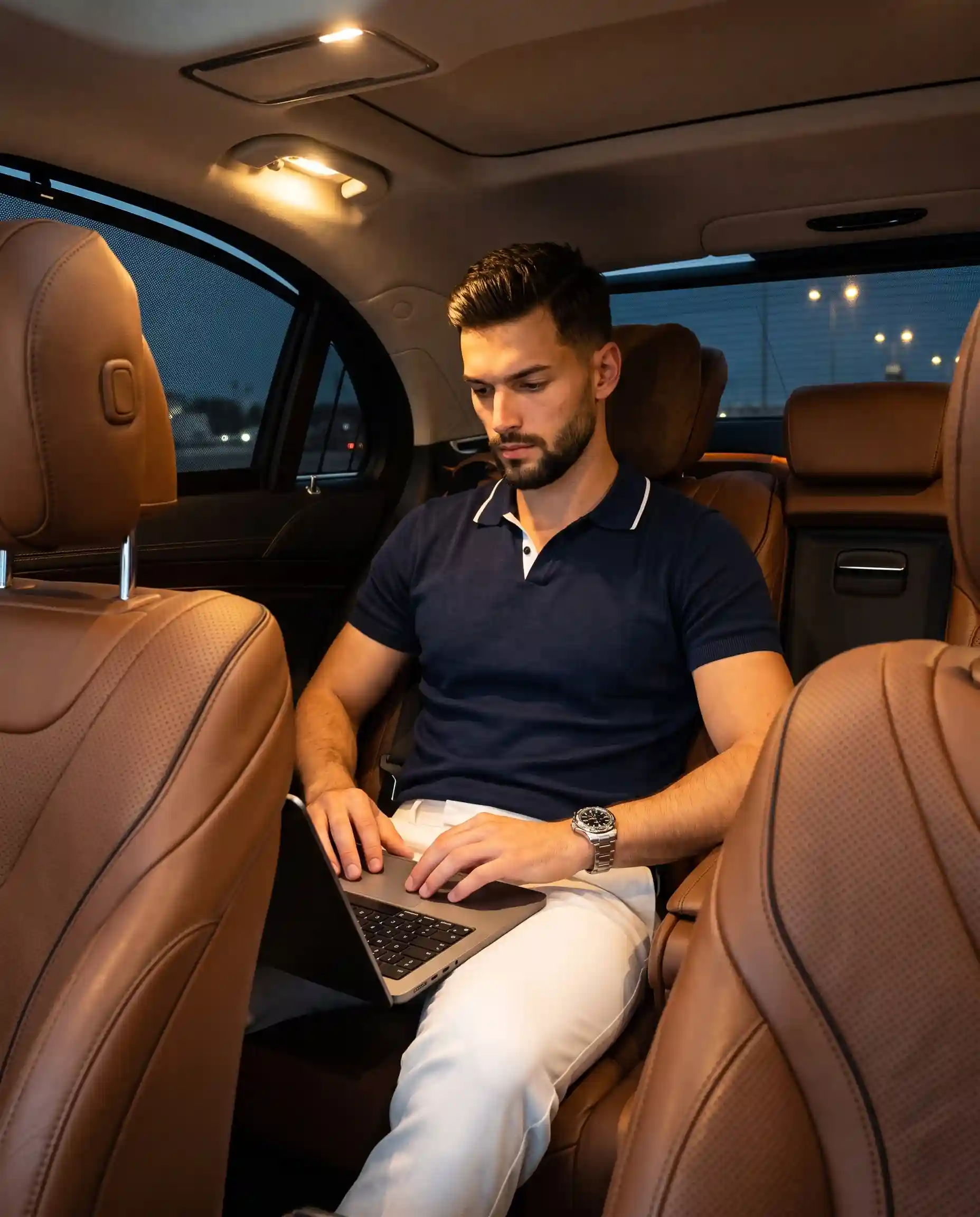 trend prompt: A handsome young adult male with messy, dark curly hair sits centered in a relaxed posture, gazing down with a serious, candid expression, closed neutral mouth, and relaxed eyebrows. He wears a dark navy polo shirt accented by a bright white-trimmed collar and crisp white trousers. A silver metal link watch adorns his left wrist, which is slightly elevated as his left hand rests casually on the left side of a silver laptop's black keyboard deck, while his relaxed right hand has its index and middle fingers lightly touching the trackpad, conveying an organic, casual readiness to type. He is positioned in the shallow spatial depth of the rear cabin of a luxury executive automobile, seated against a pristine midground of cognac brown leather featuring fine perforated patterns and precise stitched detailing. Smooth cognac brown leather front seat headrests in the foreground symmetrically flank and frame his silhouette, while a dark tinted car window to the left reflects faint exterior street lights and a subtle hint of a red seatbelt buckle edge is visible near the lower right against the black carpet. The moody, sophisticated scene is illuminated by hard, dramatic top-down lighting emanating from two warm golden overhead cabin reading lights, casting high-contrast, deep black, harshly defined shadows under his chin, beneath the laptop, and flanking the car's interior. Brilliant specular highlights are preserved on his brightly lit face, the crisp white collar, the metallic watch face, and the top edges of the luxurious leather seats, enhancing a rich complementary palette of deep navy, cognac brown, stark black, and warm white. This editorial luxury lifestyle photograph is captured digitally with tack-sharp realism and a straight-on perspective, evoking the aesthetic of a full-frame camera using a 35mm lens at f/2.0 for a medium depth of field. Shot with a high ISO of 3200 to introduce authentic digital noise, the image features cinematic post-processing with crushed blacks for dense shadow depth, enhanced color contrast between the navy shirt and brown leather, and a clarity boost on the facial features, all meticulously framed in a 4:5 aspect ratio.