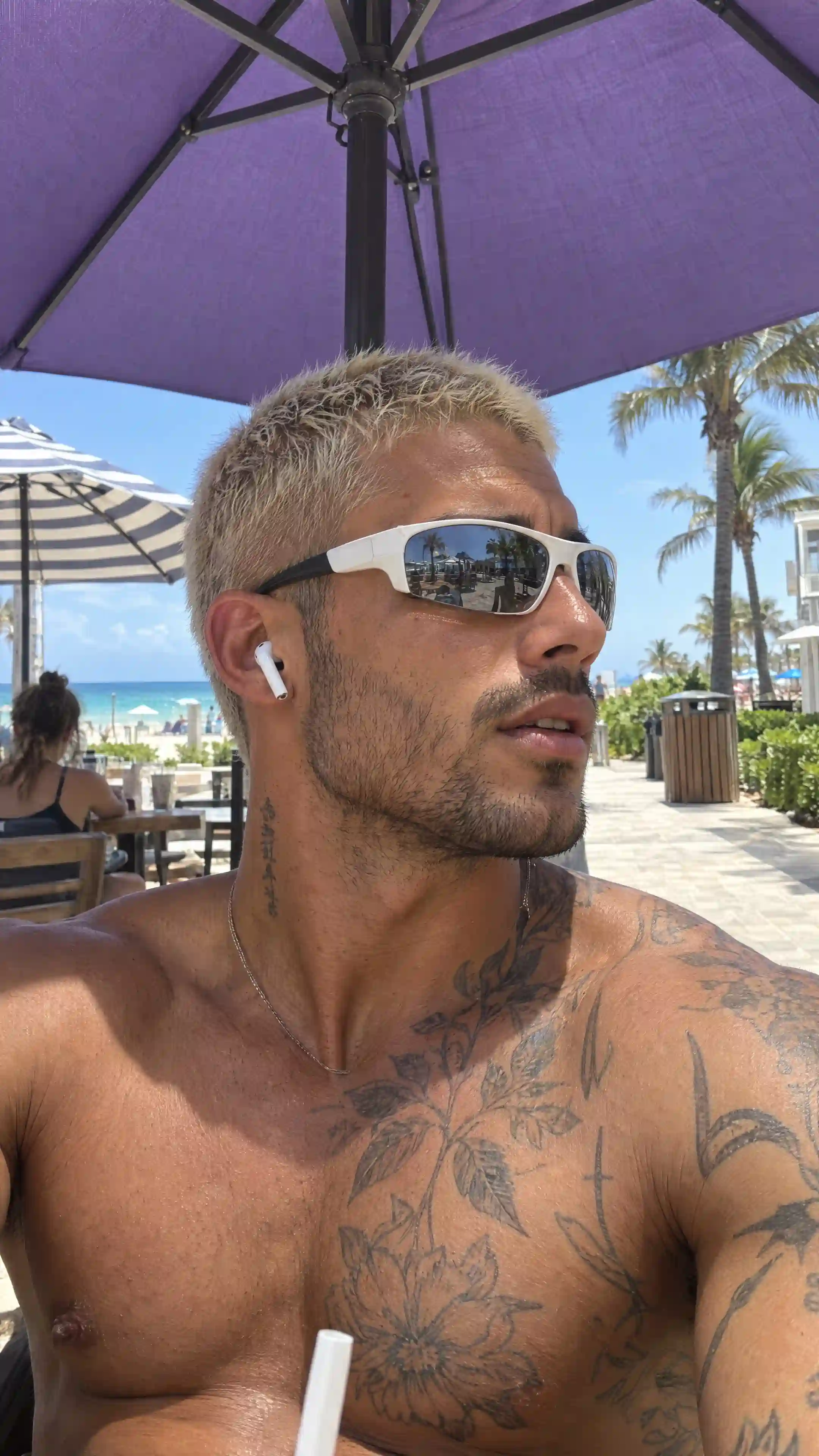 trend prompt: {"shot_type":"Vertical smartphone selfie-style portrait, casual beach lifestyle aesthetic","canvas":{"orientation":"portrait","aspect_ratio":"9:16","framing":"Close medium portrait crop from top of head to mid-chest","subject_alignment":"Centered, angled three-quarter profile","perspective":"Natural wide-angle smartphone front-camera perspective, taken from a slightly low angle looking up"},"scene":{"location":"Outdoor beachside café or boardwalk seating area","time_of_day":"Midday, bright direct sunlight","mood":"Casual, relaxed, sun-drenched beach atmosphere","environment":{"canopy":"Large purple canvas umbrella with dark metal ribs directly overhead, partially cropping the top of frame","background_left":"A second striped umbrella and a wooden café table where another person sits, blurred and out of focus","background_right":"Palm trees, a wooden trash receptacle, distant beach chairs, and a paved stone walkway leading toward the shoreline","sky":"Bright hazy blue sky visible in small gaps between the umbrella and buildings","ocean":"A thin strip of turquoise ocean and pale sand visible far in the background on the left side"}},"subject":{"description":"Young man in his late 20s, angled in profile with his face turned to the right, looking off toward the middle distance","position":"Positioned lower-center in frame, shoulders and upper chest filling the lower half","pose":{"head":"Turned in three-quarter profile, chin slightly lifted","shoulders":"One shoulder raised slightly closer to the camera due to the selfie angle","posture":"Relaxed, seated"},"expression":{"overall":"Calm, composed, slightly focused","mouth":"Lips slightly parted as if mid-breath or mid-sentence","gaze":"Directed off to the side, not toward the camera","emotion":"Relaxed confidence"},"skin":{"tone":"Deeply tanned olive-bronze skin","finish":"Natural matte-to-slight sheen typical of sun exposure and light perspiration","texture":"Realistic visible pores, natural fine lines, light natural sheen across the forehead and nose bridge","highlights":"Bright sunlit highlight along the cheekbone, nose, and shoulder"},"face":{"shape":"Angular with a strong defined jawline","cheekbones":"High and prominent","jaw":"Sharp, squared","brow":"Slightly furrowed, faint natural forehead lines visible from the angle","facial_hair":"Short, dense, well-groomed dark stubble covering the jaw, chin, and upper lip, slightly thicker along the mustache and chin","nose":"Straight, moderately defined bridge","lips":"Medium fullness, natural warm pink-brown tone, slightly parted","ear":"Visible in profile, small silver hoop-style hardware not present, ear otherwise bare aside from the earbud"}},"hair":{"color":"Bleached platinum-blonde with a slightly warm ashy undertone","length":"Very short buzzed crop","texture":"Densely textured, slightly spiky natural texture typical of short bleached hair","part":"No visible part, uniform short length across the scalp","roots":"Faint natural darker regrowth shadow near the scalp","styling":"Natural, unstyled, slightly tousled from wind and heat"},"eyewear":{"type":"Wraparound sport sunglasses","frame_color":"White frame with black temple arms","lens_color":"Dark mirrored silver-grey lenses","fit":"Wrapped closely around the temples, resting on the bridge of the nose","reflection":"Faint reflection of the surrounding beach environment visible in the lens surface","branding":"No visible brand text or logo"},"accessories":{"earbuds":"Single white wireless earbud with a visible stem, worn in the ear closest to the camera","necklace":"Thin silver chain necklace resting against the collarbone, mostly obscured by the tattoo","drink":"A white drinking straw visible at the very bottom edge of frame, held near chest height"},"tattoos":{"chest_and_shoulder":"Large detailed black ink tattoo covering the chest and shoulder, featuring botanical vine and leaf linework, a stylized flower design lower on the chest, and additional abstract linework extending toward the shoulder","neck":"A short vertical script-style tattoo visible on the side of the neck near the collarbone","style":"Fine linework, moderate density, natural fading suggesting the tattoo has some age"},"body":{"build":"Lean and muscular","visible_area":"Bare shoulders, chest, and upper torso","skin_detail":"Fine natural sheen from heat and light perspiration, visible muscle definition across the shoulder and chest"},"lighting":{"type":"Bright natural harsh midday sunlight","primary_source":"Direct overhead sun, partially filtered by the umbrella canopy","direction":"From above and slightly behind, casting a defined shadow line across part of the face and neck","quality":"High contrast between sunlit and shaded areas","highlights":"Strong specular highlight on the cheekbone, nose bridge, and shoulder","shadow_style":"Hard-edged natural shadow along the jaw and neck from the umbrella's edge","color_temperature":"Warm daylight with slightly cool ambient fill from the surrounding sky and umbrella shade"},"style":"Ultra-realistic, photorealistic, authentic candid smartphone selfie aesthetic, natural skin texture with visible pores and sheen, no digital smoothing, no plastic-looking skin","aspect_ratio":"9:16","quality":"high detail, realistic skin, hair, and tattoo texture, natural color grading, no exaggerated anatomy"}
