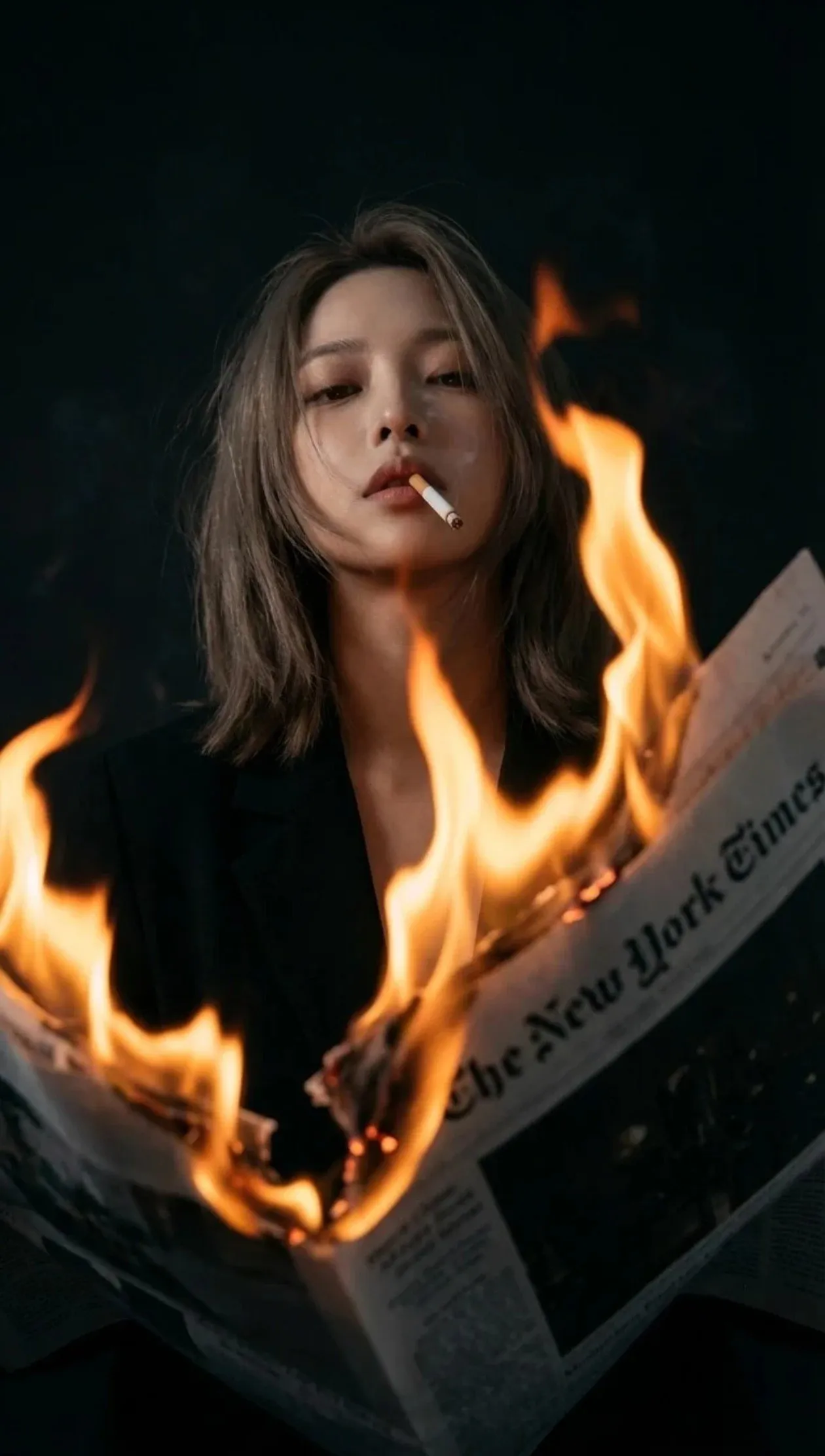 trend prompt: A young woman with slightly tousled shoulder-length hair, cigarette in mouth, viewed from BELOW through flames. Shoulder-length hair with natural slight waves, a little tousled but not wild - effortlessly messy, lived-in texture. Looking down toward camera through the fire, half-lidded distant gaze. Cold, detached, nonchalant expression with cigarette dangling from lips. Holding burning newspaper, only lower face visible - chin, lips, cigarette, and jaw seen through flames. Black blazer, dark formal wear. The New York Times newspaper held in front, bottom edge of newspaper and flames are in EXTREME FOREGROUND, burning with flames rising UP toward camera. FOREGROUND ELEMENT - camera shoots THROUGH the flames, fire partially obscures and frames the view of subject's lower face. Fire between camera and subject's face, flames rising in front of lens. Warm orange and golden yellow flames, slightly out of focus in foreground. Fire creates natural vignette/frame, viewer looks THROUGH flames to see chin, lips, and cigarette. Cigarette dangling from lips, lit, visible through the fire, smoke mixing with fire glow. Close-up of lower face THROUGH foreground flames, 35mm f/1.8. LOW ANGLE from below newspaper level, shooting UP through fire toward chin/mouth. Camera placed as if BEHIND the burning newspaper edge, shooting through flames. FG: out-of-focus fire and newspaper edge filling bottom/sides of frame; MG: sharp focus on chin, lips, cigarette; BG: dark face fading into black. Shallow depth of field - foreground flames slightly blurred, focus on lips/chin area. SINGLE LIGHT SOURCE - fire only. NO other light sources, NO fill light, NO ambient light. ONLY the burning newspaper flames illuminate the scene. Fire below face, casting warm uplight on chin, underlip, and nose from below. Dark, dramatic, firelit chiaroscuro. PURE BLACK surroundings, face emerges from complete darkness, lit only by fire glow from below. Fujifilm ETERNA style, extremely dark and underexposed, fire is the ONLY color/light in the frame. Pure black background, warm orange fire glow on skin, deep shadows everywhere else. Very dark overall exposure with crushed blacks. Intimate yet menacing, voyeuristic through flames, dark feminine power.