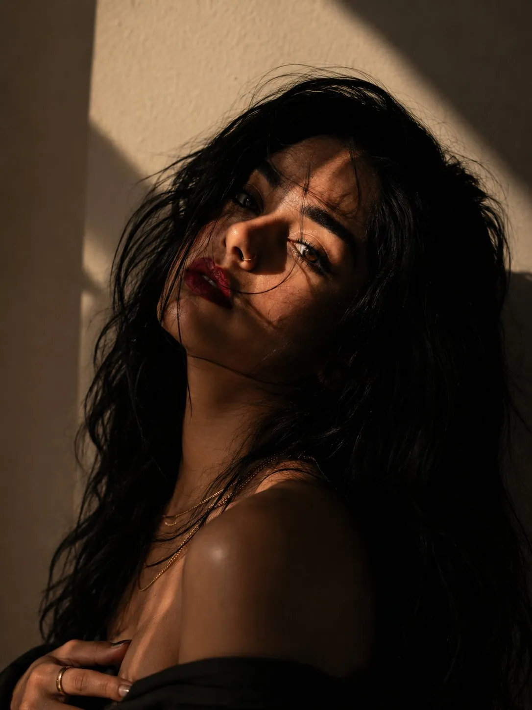 trend prompt: Create an ultra-realistic, cinematic fine-art portrait of a beautiful young woman with a moody, editorial aesthetic. She is shown in a close-up head-and-shoulders composition, gently tilting her head to one side while looking directly into the camera with a calm, mysterious, and emotionally captivating expression. She has long, naturally wavy black hair, with a few loose strands softly blowing across her face. Her skin is smooth with natural texture, thick dark eyebrows, long eyelashes, and deep brown eyes that catch the light beautifully. She wears soft matte deep-red lipstick, minimal natural makeup, a small silver nose ring, and layered delicate gold necklaces, creating an effortlessly elegant look. Her shoulders are bare for a timeless editorial portrait style. A dramatic band of golden window light falls diagonally across one side of her face, while the remaining areas fade into rich, soft shadows. The lighting creates striking contrast, adding depth, mystery, and cinematic emotion. The background is a simple warm off-white textured wall with no distractions. Composition: close-up portrait, slight Dutch-angle head tilt, centered framing, shallow depth of field, editorial beauty photography, luxury fashion magazine style, powerful eye contact, minimalist composition, emotionally expressive, stop-scrolling, premium Instagram portrait. Lighting: warm natural sunlight through window blinds, high contrast, cinematic chiaroscuro, soft shadow transitions, realistic skin highlights, subtle ambient fill light. Camera: Sony A7R V, FE 85mm f/1.4 GM II, f/1.4, ISO 100, HDR, RAW, ultra-sharp focus on the eyes, creamy background blur, natural color grading, DSLR-quality, 8K, photorealistic, highly detailed, no AI artifacts, no oversmoothing, realistic skin pores and fine hair strands. Aspect Ratio: 3:4.
