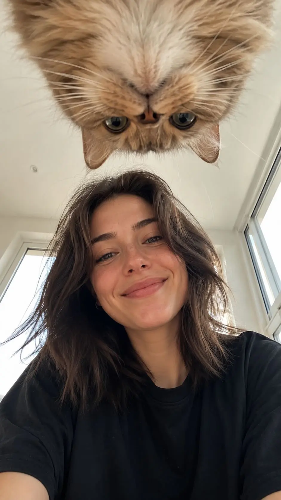 trend prompt: Create a realistic vertical smartphone selfie taken indoors from a very low floor-level angle, with the camera pointing upward. A woman with fair skin, naturally proportioned facial features, expressive eyes, and shoulder-length dark hair stands in the lower center of the frame wearing an oversized black T-shirt. She looks down into the camera with a warm genuine smile, bright friendly eyes, and a relaxed social-media selfie expression. At the top center of the frame, an upside-down fluffy cat leans toward the lens, looking directly downward and dominating the upper composition with a cute, funny, and charming expression that matches the woman's friendly and cheerful mood. Bright modern apartment interior, large windows, soft natural daylight, clean minimal background. Ultra-realistic skin rendering with visible natural pores, fine facial hairs, subtle skin texture, realistic eyelids, natural lip texture, soft under-eye details, authentic complexion variations, slight redness in natural areas of the face, realistic skin translucency, no beauty filter effect, no plastic skin, no excessive retouching, realistic facial depth and micro-details. Matte skin finish with natural light absorption, true-to-life skin tones and highly realistic texture reproduction. Professional portrait photography quality captured with a Sony A7R V, 85mm f/1.8 portrait lens, f/2.0 aperture, ISO 100, ultra-high-resolution sensor, shallow depth of field, natural optical bokeh, realistic lens rendering, high dynamic range, accurate skin color reproduction, crisp focus on the face and eyes. Soft window light combined with realistic ambient indoor illumination, natural shadow transitions, subtle catchlights in the eyes, cinematic yet realistic lighting. Highly detailed cat fur, individual hair strands visible, realistic whiskers and facial features. Natural smartphone-photo composition with balanced framing, candid atmosphere, social-media-friendly aesthetic, photorealistic, ultra-detailed, lifelike textures, realistic lighting physics, premium portrait quality, 8K resolution. Negative prompt: duck face, serious expression, eye-level camera, flat selfie angle, side-positioned animal, random animal, cartoon, anime, CGI, artificial skin smoothing, plastic skin, beauty filter, glamour retouching, waxy skin, distorted anatomy, extra limbs, messy room, scary expressions, aggressive animal behavior, blur, low resolution, watermark, logo, text.