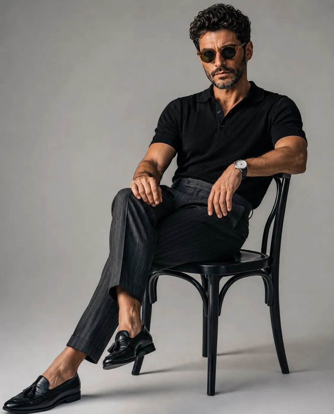 trend prompt: A luxury editorial fashion portrait of a stylish young man sitting casually on a classic black wooden chair in a minimalist photography studio with a seamless light gray backdrop. He wears a fitted black knit polo shirt tucked into tailored high-waisted charcoal gray pinstripe trousers, paired with polished black leather tassel loafers and a minimalist silver wristwatch. One leg is extended while the other is crossed naturally, with one arm resting on his knee and the other relaxed on the chair, maintaining direct eye contact with a calm, confident expression. Slightly messy textured hair, clean masculine features, relaxed yet elegant posture. Soft diffused studio lighting from a large octabox creates gentle shadows, realistic skin tones, and subtle depth. Minimalist composition with negative space, quiet luxury aesthetic, old-money fashion, premium menswear editorial, ultra-realistic, photorealistic, Vogue magazine style, Hasselblad medium format look, 85mm lens, f/4, crisp details, cinematic color grading, high dynamic range, 8K. Negative Prompt: low quality, blurry, cartoon, anime, CGI, unrealistic anatomy, extra fingers, duplicate limbs, distorted face, bad proportions, oversaturated colors, harsh lighting, watermark, logo, text, cluttered background, noise, artifacts, motion blur, low resolution, overexposed, underexposed, wrinkled clothing, awkward pose.