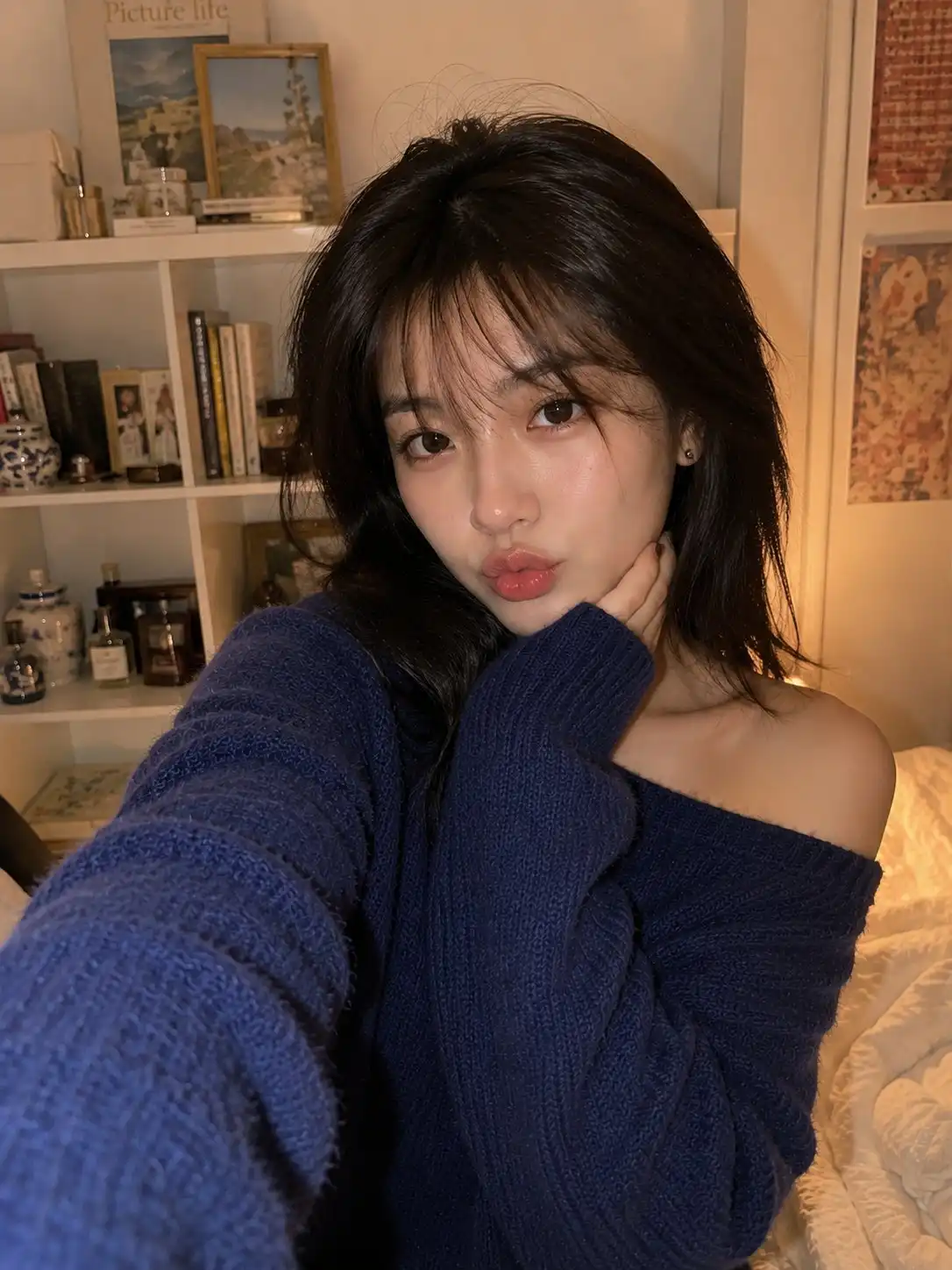 trend prompt: Smartphone selfie of a beautiful young East Asian woman indoors in a cozy bedroom. She has porcelain-fair skin, large expressive dark brown eyes, naturally full lips, a small straight nose, and soft oval facial features. Her medium-length dark brown hair is slightly messy with airy wispy bangs falling naturally across her forehead, giving a fresh just-woke-up look. She is making a subtle duck-lips expression while looking directly into the phone camera. She wears an oversized fuzzy knitted navy-blue off-shoulder sweater that casually slips off one shoulder. The background features a white bookshelf filled with books, candles, small decorative jars, framed artwork, and cute vintage-style room decor, creating a warm, aesthetic atmosphere. Captured casually with a modern smartphone at arm's length, eye-level angle, vertical portrait orientation. Natural indoor ambient lighting mixed with warm room light, soft shadows, realistic skin texture, slight smartphone HDR processing, tiny sensor noise, subtle lens distortion from a 26mm equivalent mobile camera, automatic white balance, authentic exposure, casual composition, slightly imperfect framing, no professional lighting, no beauty filter, no AI look, everyday social media selfie, ultra-photorealistic.