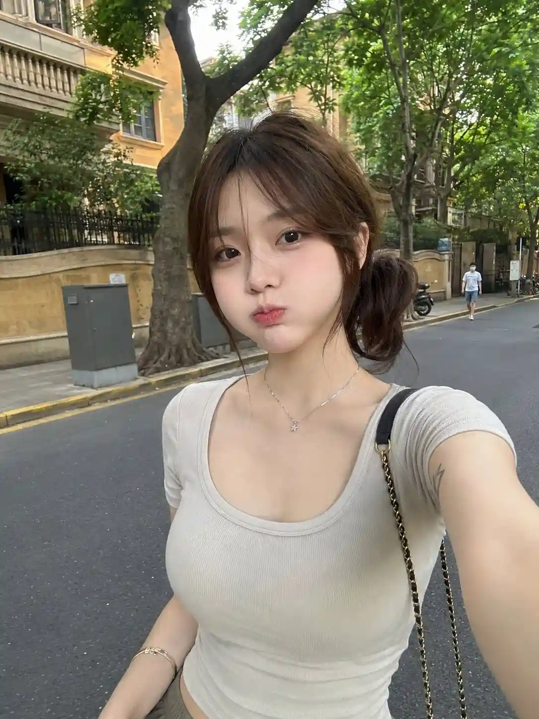 trend prompt: A RAW, ultra-realistic smartphone photo of a beautiful young East Asian woman standing casually on a quiet tree-lined city street during the daytime. She has fair porcelain skin, large expressive dark brown eyes, a small straight nose, naturally soft pink lips, and delicate youthful facial features. Her medium-length chestnut brown hair is tied into a low side ponytail with soft wispy curtain bangs framing her face. She is making a subtle puffed-cheek expression while looking directly into the camera with a playful yet natural look. She wears a fitted light beige scoop-neck short-sleeve T-shirt that hugs her silhouette naturally, a delicate silver necklace with a tiny star pendant, a thin gold bracelet, and carries a small black leather shoulder bag with a gold chain strap. A small minimalist line-art tattoo is visible on her upper arm. The background features a peaceful urban street with lush green trees, elegant residential buildings, a quiet sidewalk, parked scooters, and a pedestrian walking in the distance, creating a realistic everyday city atmosphere. Captured casually by a friend using a modern smartphone from a standing eye-level angle. Natural late-afternoon daylight, soft diffused lighting, realistic skin texture, subtle smartphone HDR processing, tiny sensor noise, automatic white balance, authentic exposure, slight 26mm equivalent mobile lens perspective distortion, casual composition, slightly imperfect framing, no professional lighting, no beauty filters, no AI-generated appearance, everyday lifestyle photography, ultra-photorealistic, RAW smartphone camera quality, 8K.