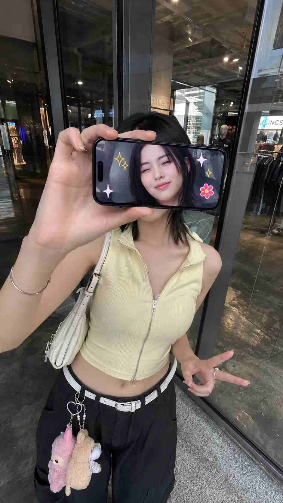 GPT Image 2 prompt: Ultra-realistic creative portrait taken with an iPhone, identity accurately preserved from the reference image. A woman stands inside a store, facing a glass display window or a reflective wall, photographed from a slightly elevated frontal angle. She holds a smartphone horizontally in front of her face, covering her eyes and the upper part of her face. The phone's screen points at the camera and clearly displays a real-time image of her face.