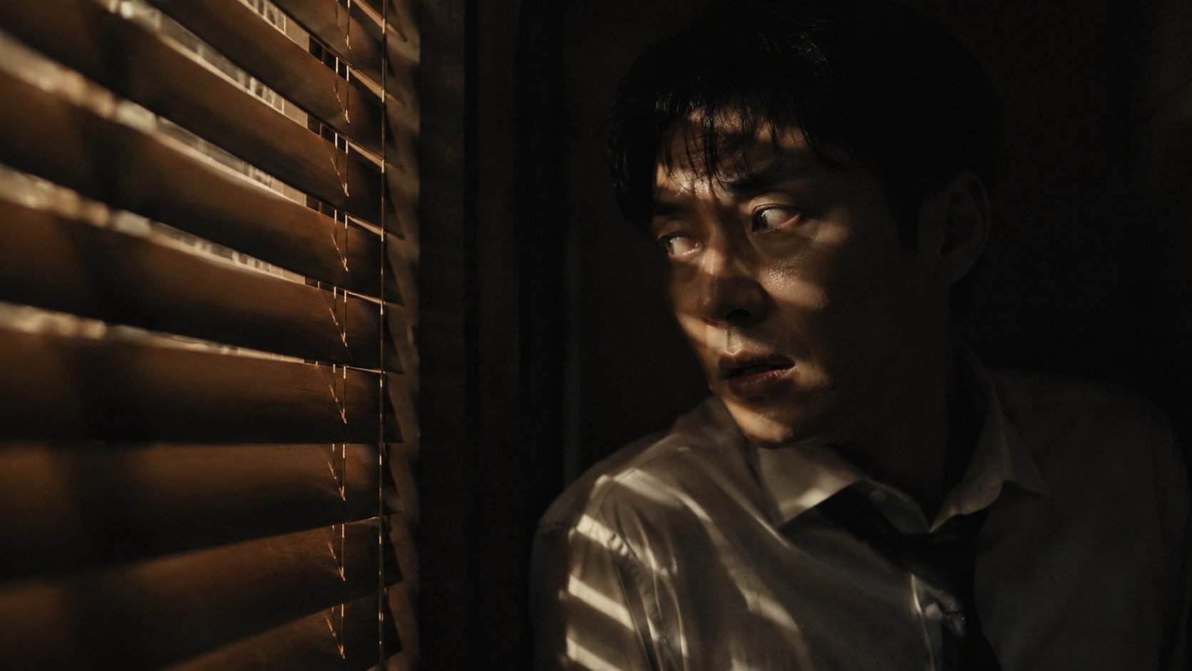 GPT Image 2 prompt: Create a dark cinematic thriller film still of an East Asian man hiding in a narrow shadowy space beside wooden blinds or shutters. He is partially hidden in darkness, wearing a white shirt with a loosened dark tie, his face tense and alert. Thin strips of warm light pass through the blinds, cutting across his face and body, revealing only his eyes, cheekbones, and part of his anxious expression. His mouth is slightly open as if he is breathing quietly or listening for danger nearby.

The scene should feel claustrophobic, suspenseful, and intimate, with deep black shadows surrounding him. The wooden slats create strong horizontal shadow patterns across the frame. Use low-key lighting, high contrast, warm golden highlights mixed with deep darkness, realistic facial detail, cinematic film grain, shallow depth of field, anamorphic widescreen composition, tense psychological thriller atmosphere, no text, no logo.