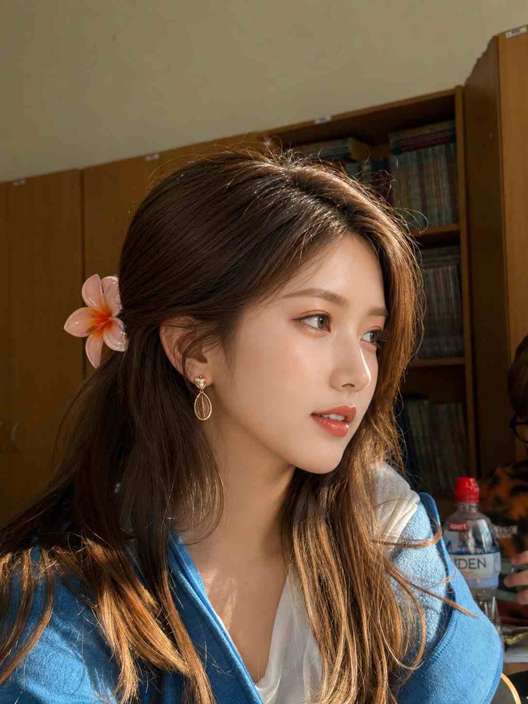 GPT Image 2 prompt: Ultra realistic aesthetic portrait of a young East Asian woman sitting in a classroom/library during golden hour sunlight, wearing a blue hoodie over a white top, soft glossy brown hair with a pink flower clipped behind her ear, elegant gold earrings, sharp facial features inspired by Korean beauty aesthetics, smooth glowing skin, natural makeup, looking sideways with a calm dreamy expression, cinematic warm lighting hitting her face, wooden bookshelves in background, shallow depth of field, ultra detailed hair strands, realistic shadows, cozy academic atmosphere, DSLR photo, 85mm lens, f/1.8, high sharpness, soft highlights, premium color grading, natural candid composition, photorealistic, 4k, clean aesthetic, balanced skin texture, subtle sunlight bloom, luxury Instagram aesthetic style