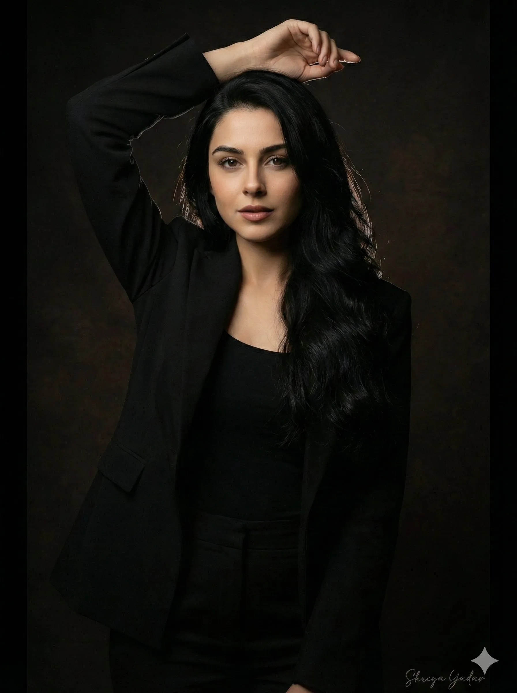 nano banana prompts: Ultra-realistic studio portrait of a woman with long wavy black hair styled to fall over one shoulder, exact face, expression, and natural skin tone. Dramatic low-key lighting with soft rim light highlighting her hair. She wears a black outfit and black blazer, posing confidently with one arm raised above her head. Dark background, sharp details, elegant and bold mood.