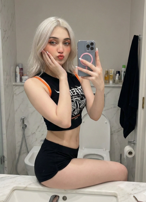 vip prompts: {
  "image_metadata": {
    "type": "Mirror selfie",
    "angle": "Straight-on frontal",
    "style": [
      "Ultra-realistic",
      "High detail",
      "Natural skin texture",
      "Seductive",
      "Casual",
      "Intimate",
      "Contemporary"
    ]
  },
  "subject": {
    "demographics": "Young woman",
    "pose": {
      "position": "Sitting on the edge of a white bathroom sink",
      "legs": "Slightly apart, one leg hanging down naturally",
      "head": "Resting gently on left palm",
      "hands": {
        "left": "Supporting head, fingers touching face",
        "right": "Holding smartphone at face level, slightly tilted"
      }
    },
    "facial_features": {
      "expression": "Subtle duck face, slightly pursed lips",
      "eyes": "Direct contact with the camera",
      "mood": "Playful, flirtatious, confident"
    },
    "details": {
      "nails": "Long light-colored French-style manicure",
      "physique": "Flat toned abdomen"
    }
  },
  "outfit": {
    "top": {
      "color": "Black",
      "style": "Short sleeveless crop top",
      "details": "Orange shoulder straps, white graphic/text print on chest (partially visible)"
    },
    "bottom": {
      "color": "Black",
      "style": "Very short shorts (sport or pajama style)",
      "fit": "Figure-emphasizing"
    }
  },
  "props": {
    "smartphone": {
      "model": "iPhone",
      "case": "Transparent",
      "accessory": "Pink MagSafe ring"
    }
  },
  "environment": {
    "location": "Modern bathroom",
    "surfaces": {
      "walls": "White marble with soft gray veining",
      "fixtures": "White sink, white toilet"
    },
    "background_elements": [
      "Bathtub or shower area",
      "Shelf with assorted bottles and toiletries (left)",
      "Black towel on a hook (left)",
      "Personal care items near toilet (right)"
    ]
  },
  "lighting": {
    "type": "Soft, even indoor lighting",
    ...}